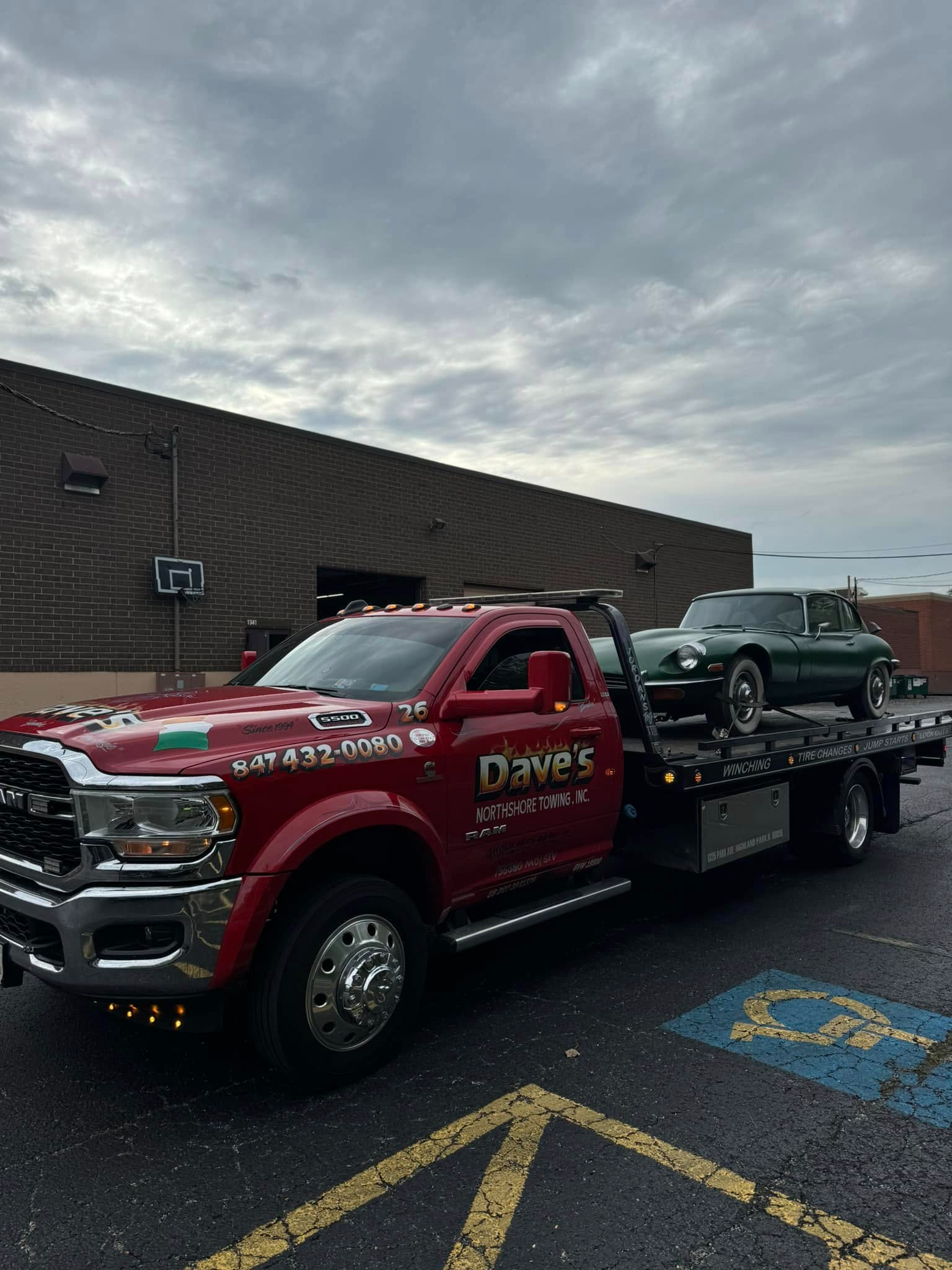 A red tow truck with a green car on the back is parked in a handicapped parking spot.