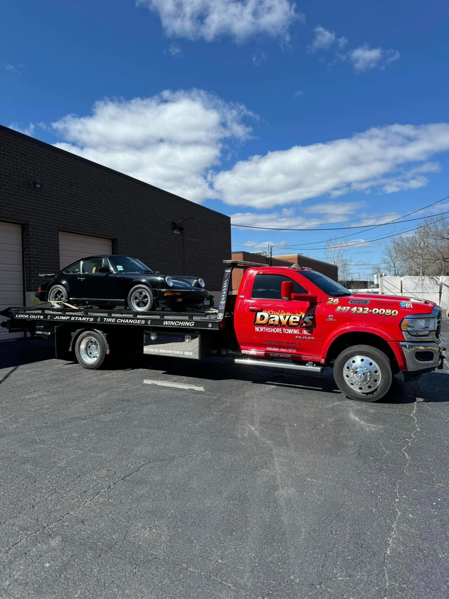 A red tow truck is towing a black car in a parking lot.