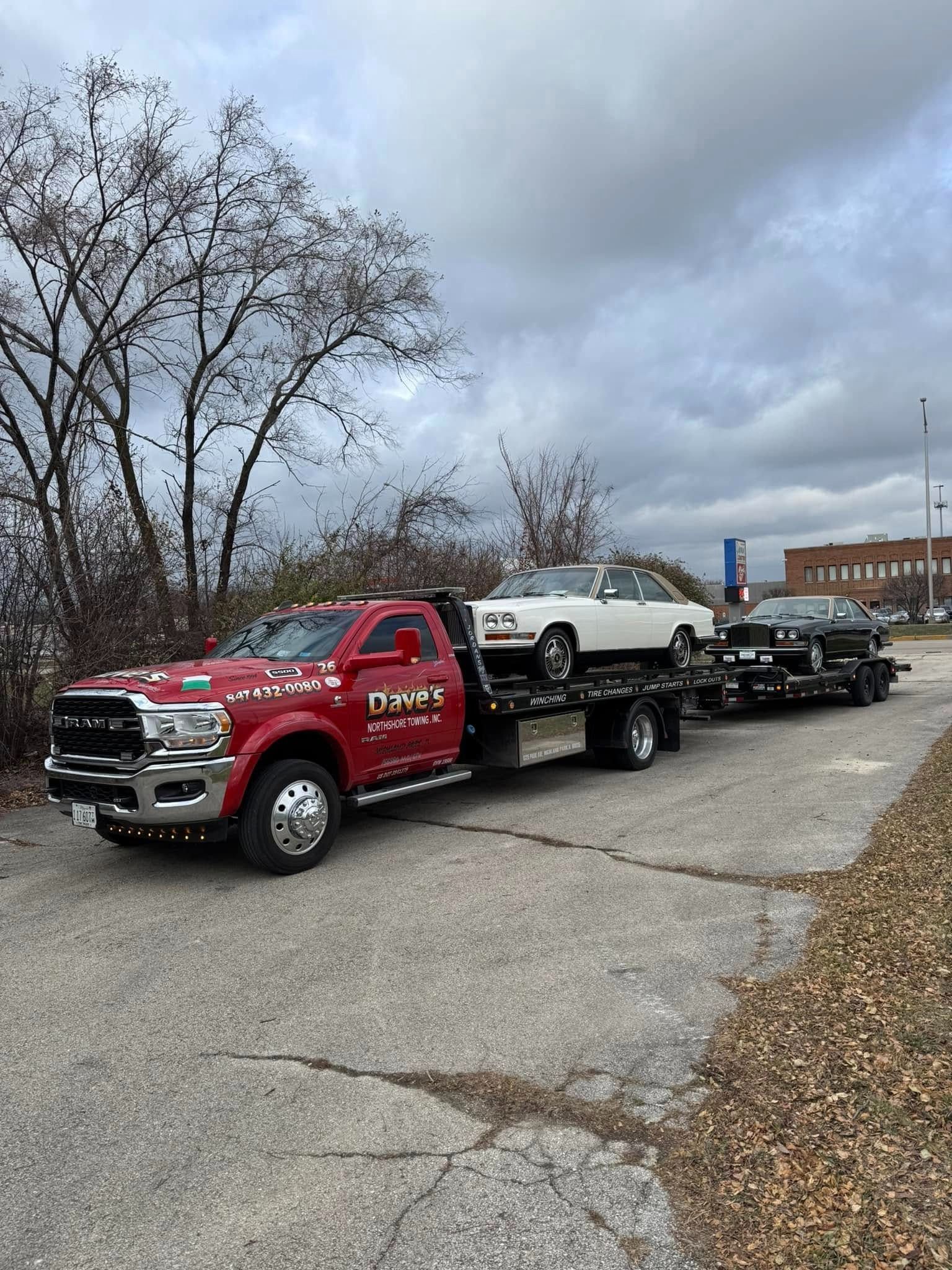 A red tow truck is carrying two cars down a road.