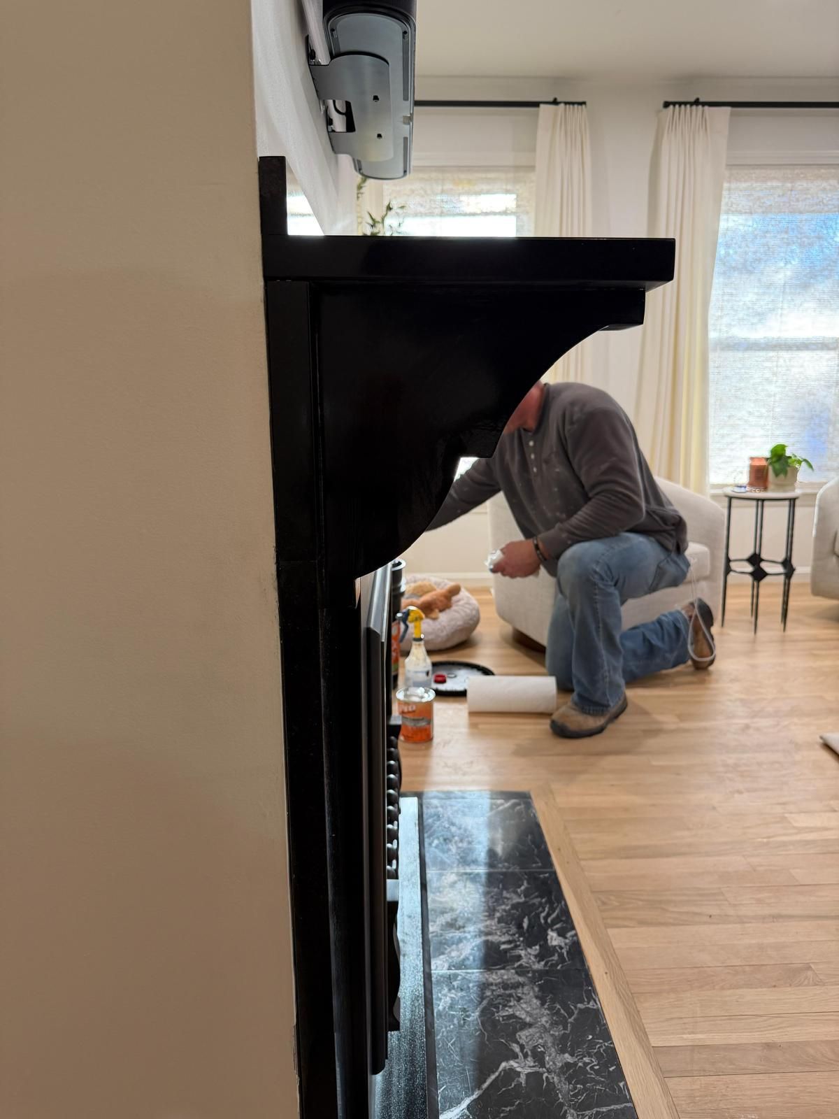 Man installing something on a black fireplace. Living room with light wood floors and sunlight coming through a window.