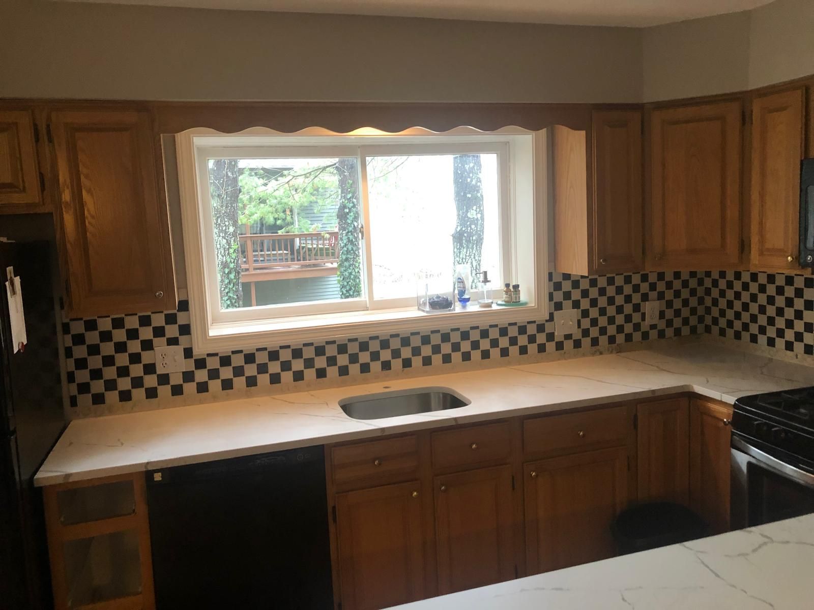 Kitchen with light wood cabinets, black and white checkered backsplash, sink, and window overlooking a deck.