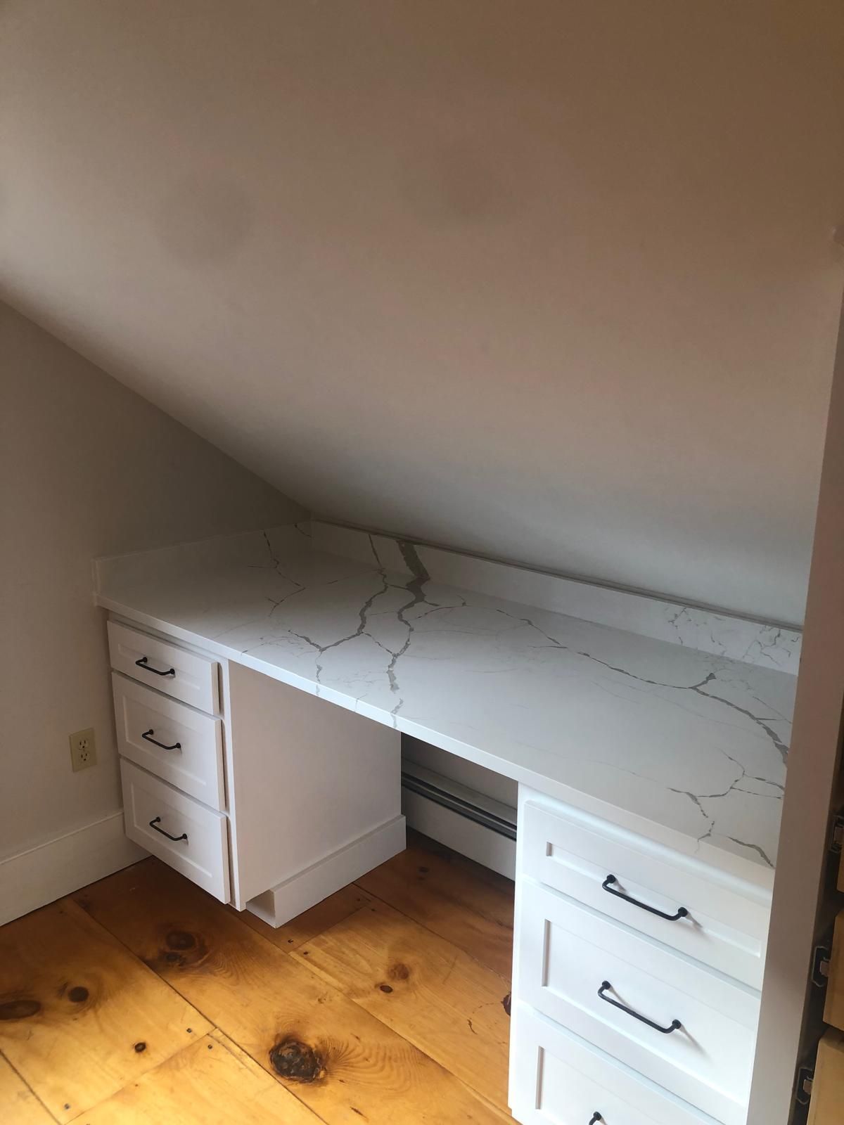 Built-in white desk with drawers, marble-patterned countertop in an attic room with wooden floor.