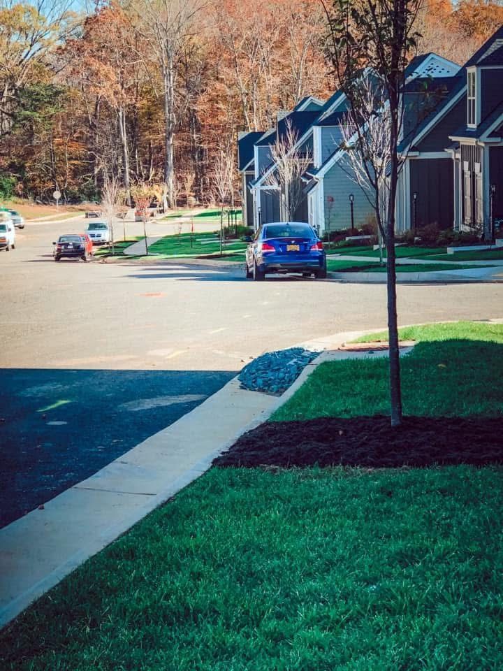 A blue car drives down a street lined with houses and fall foliage on a sunny day.