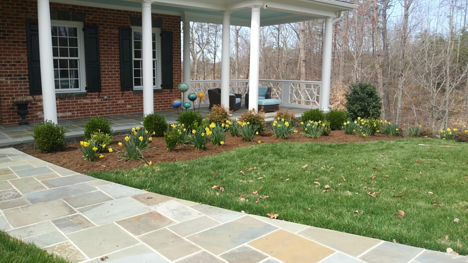 Stone walkway leading to a brick house with a porch and landscaped flower beds.