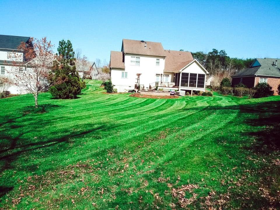 Green lawn with distinct mowing patterns, leading to a two-story beige house with a screened porch on a sunny day.