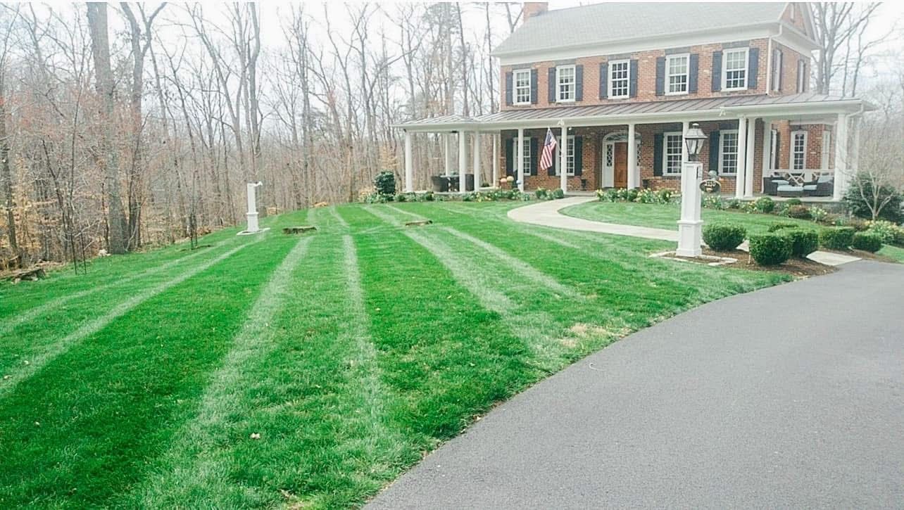 Lawn with striped pattern leading to a brick house with a porch. Trees in background, driveway on the right.