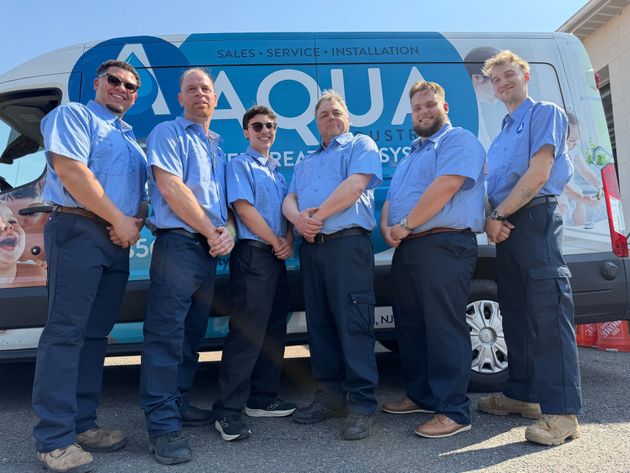 Six people wearing matching blue shirts and dark pants stand in a line before a white van with the Aqua logo.
