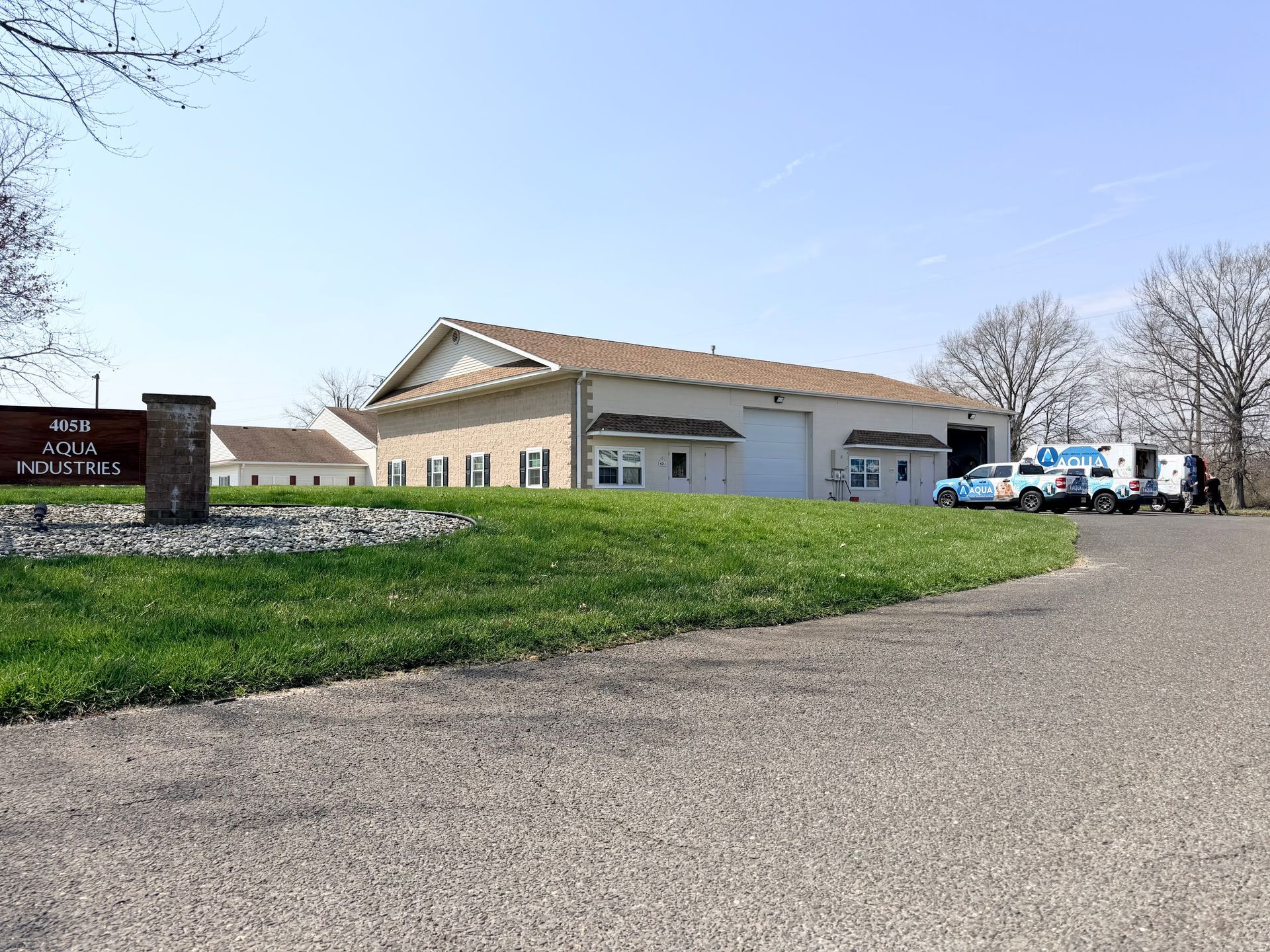 Tan commercial building with white garage doors on a grassy lot, viewed from a gravel driveway under a clear blue sky.