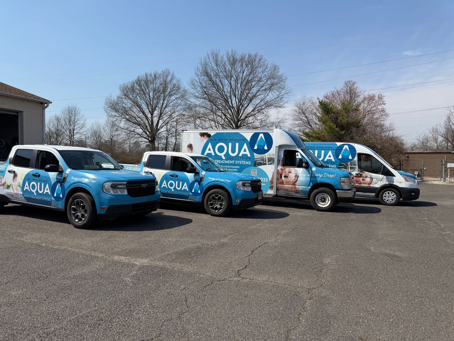 Four light-blue Aqua service vehicles, including two trucks and two vans, parked on a gravel lot under a clear blue sky.