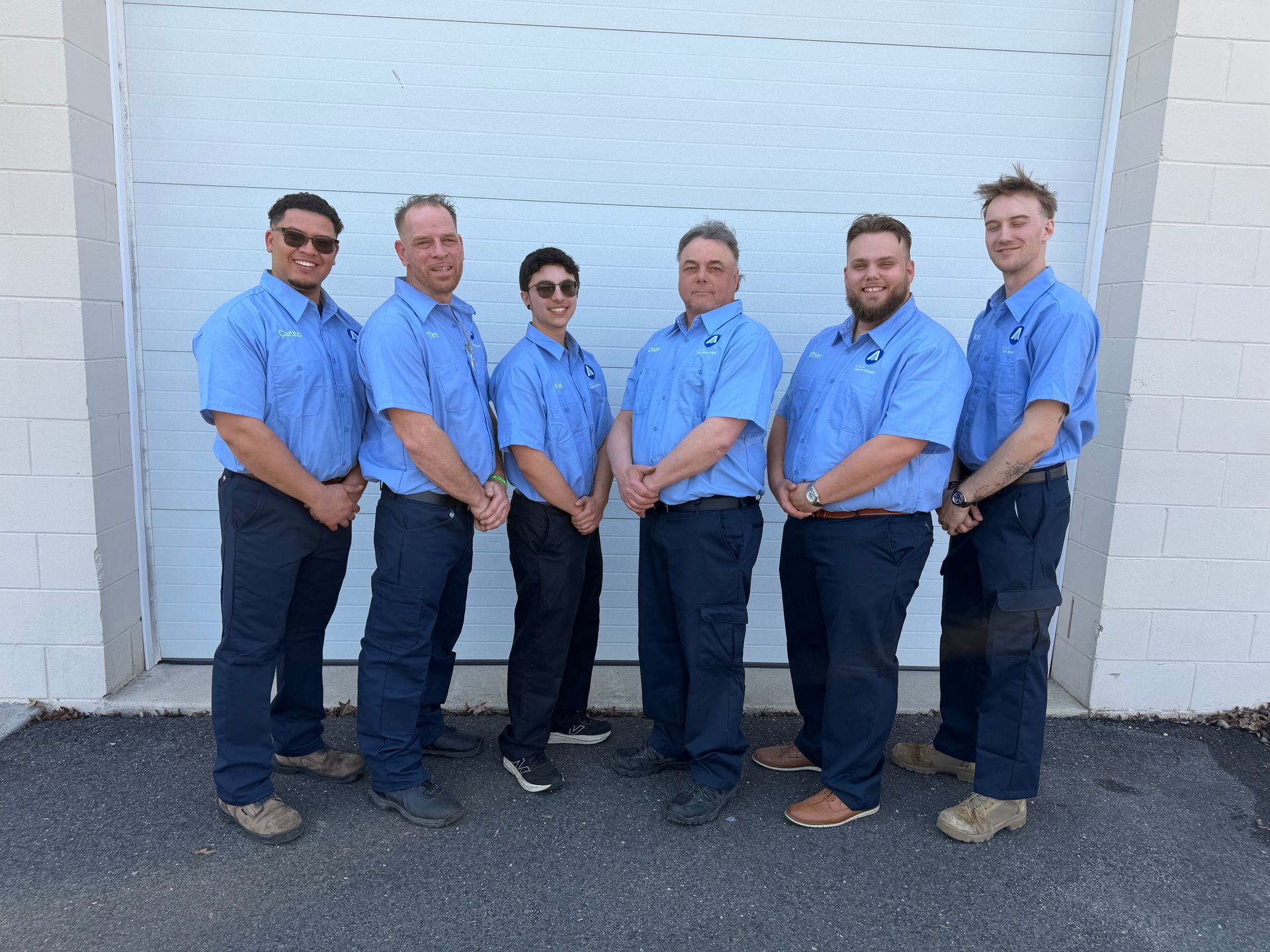 Six people in blue uniform shirts and dark pants stand in a row against a white garage door.