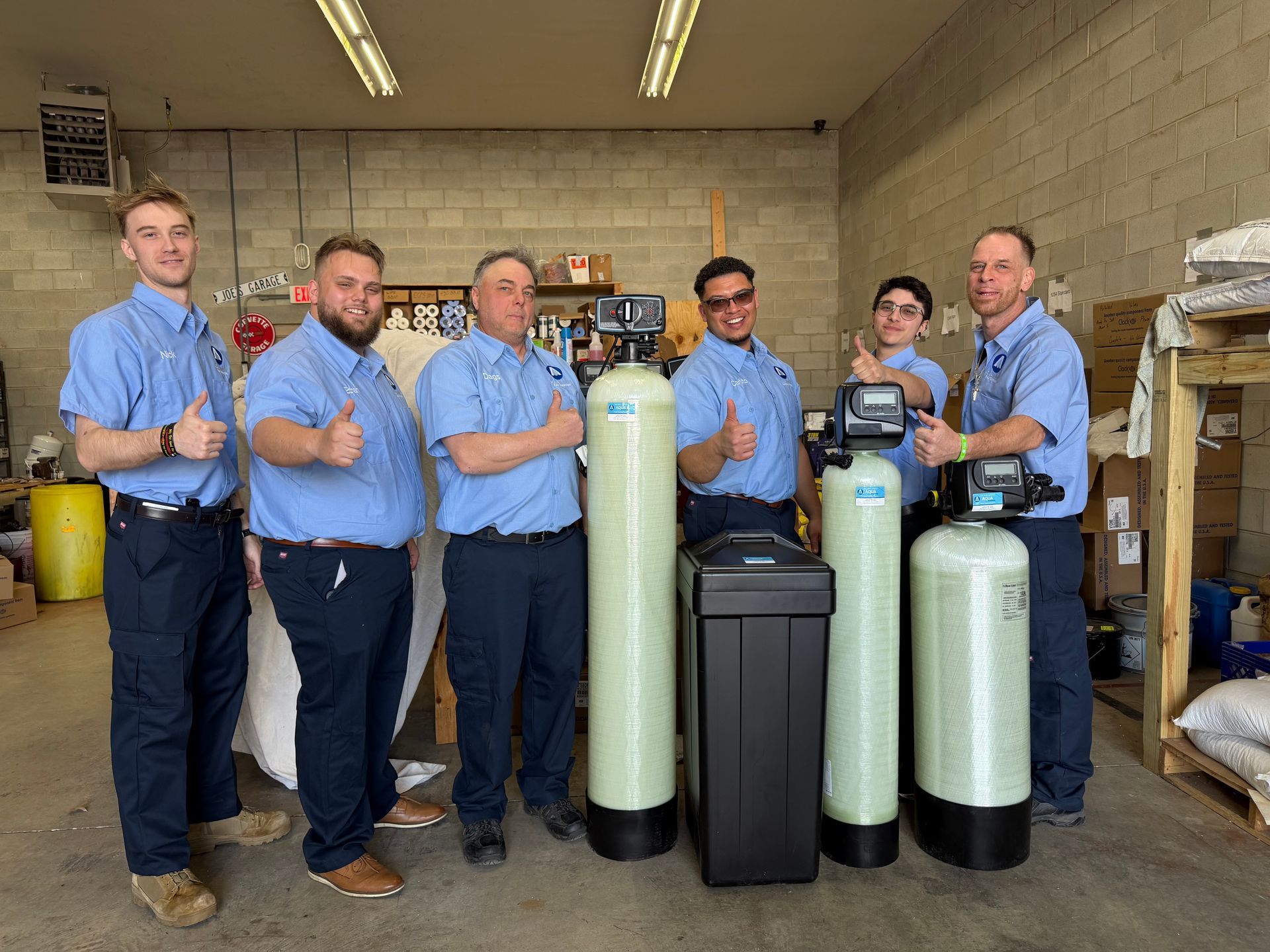 Six people in blue uniforms pose with water filtration equipment inside a warehouse, some gesturing thumbs up.