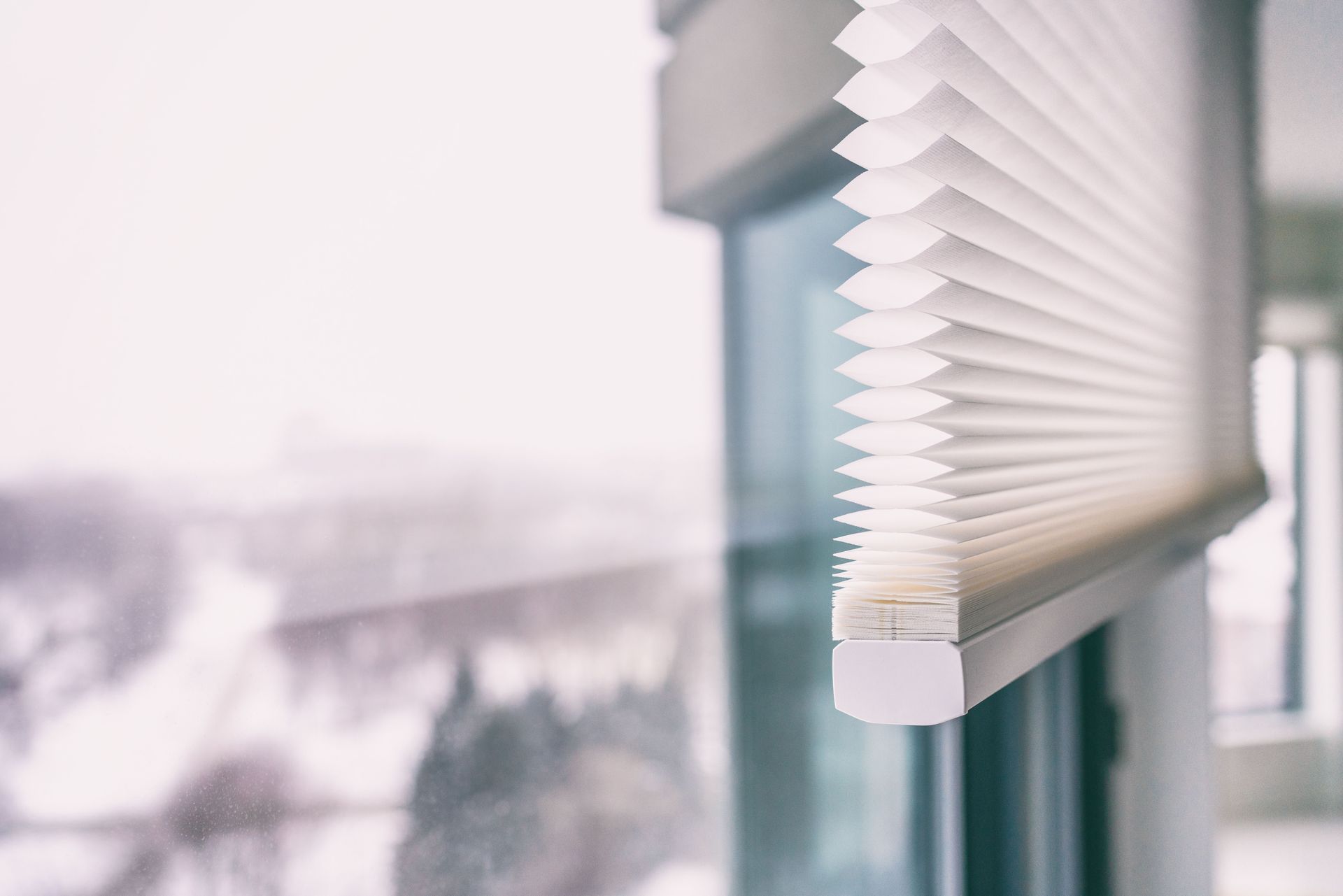 White pleated window shade partially lowered, blurred snowy outdoor view.