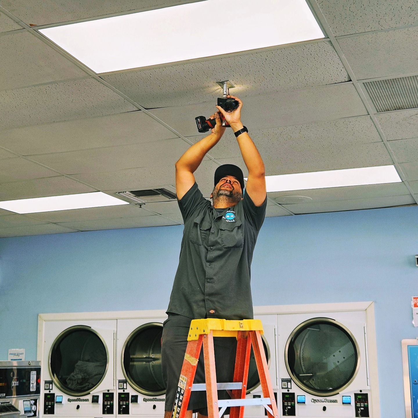 Man on stepladder installing light fixture in a laundromat, using a drill.