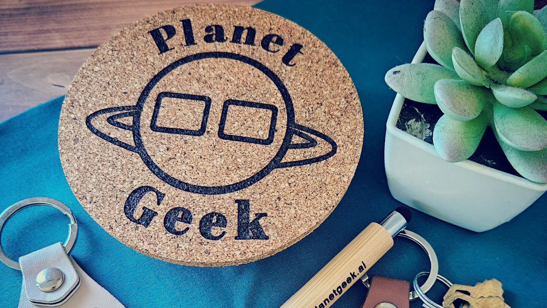 Cork coaster with "Planet Geek" logo, a succulent plant, and keychains on a blue surface.