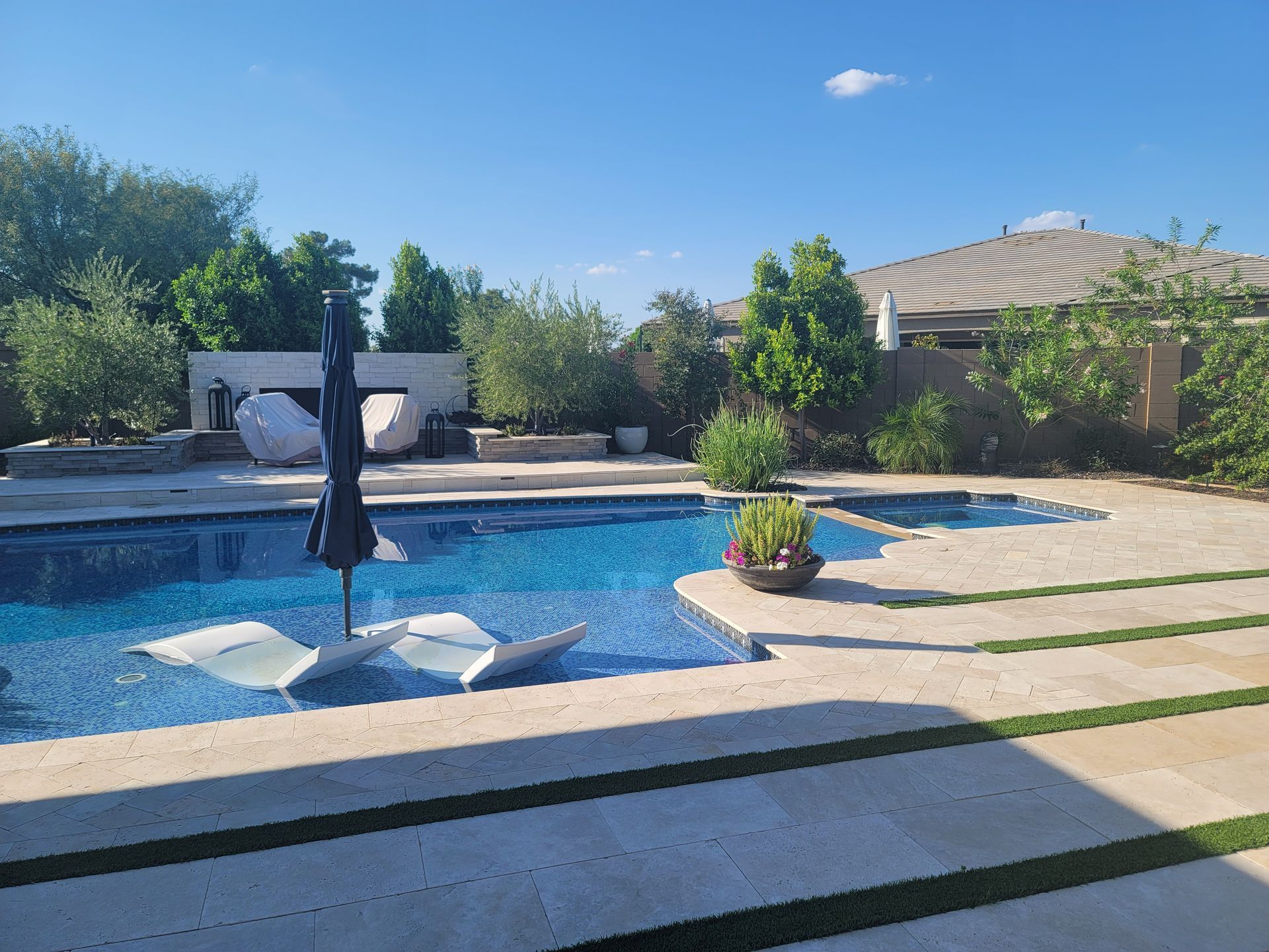 Swimming pool with floating chairs and a black umbrella, surrounded by stone patio and landscaping under a sunny sky.
