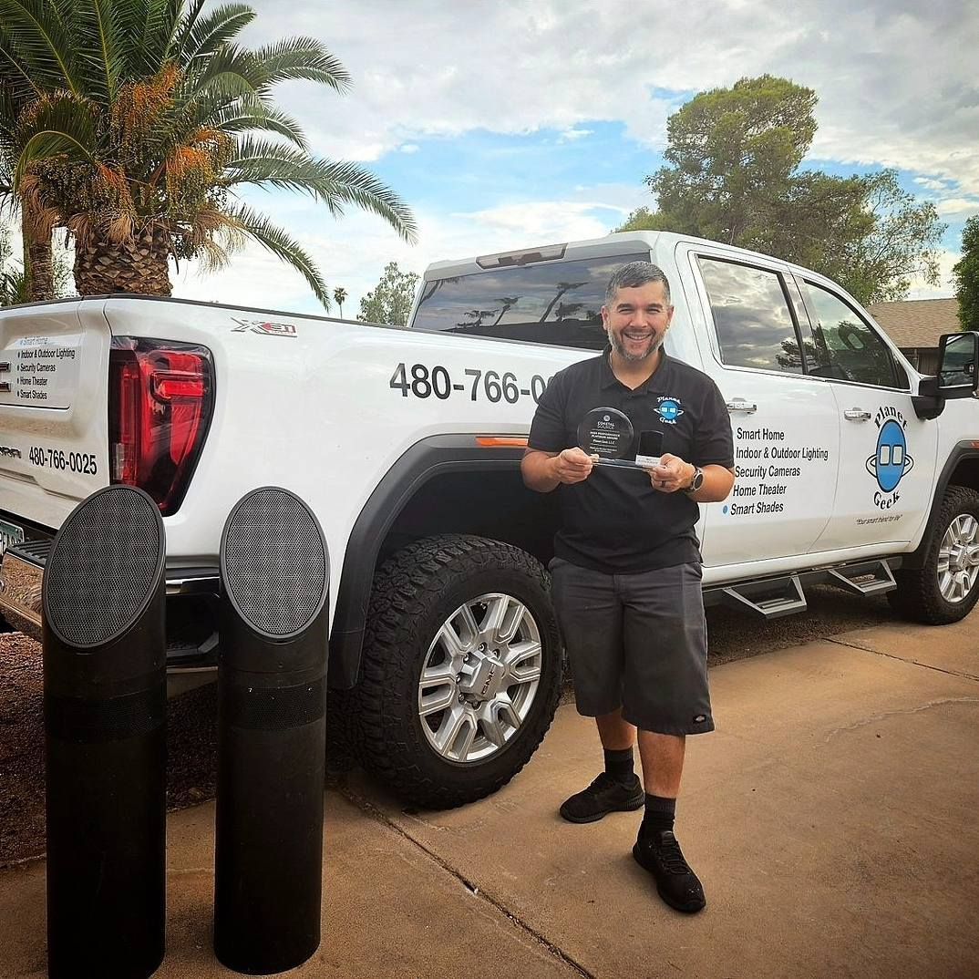 Man stands by truck, holding award. Two black cylindrical speakers on the left. White truck with logo in the background.