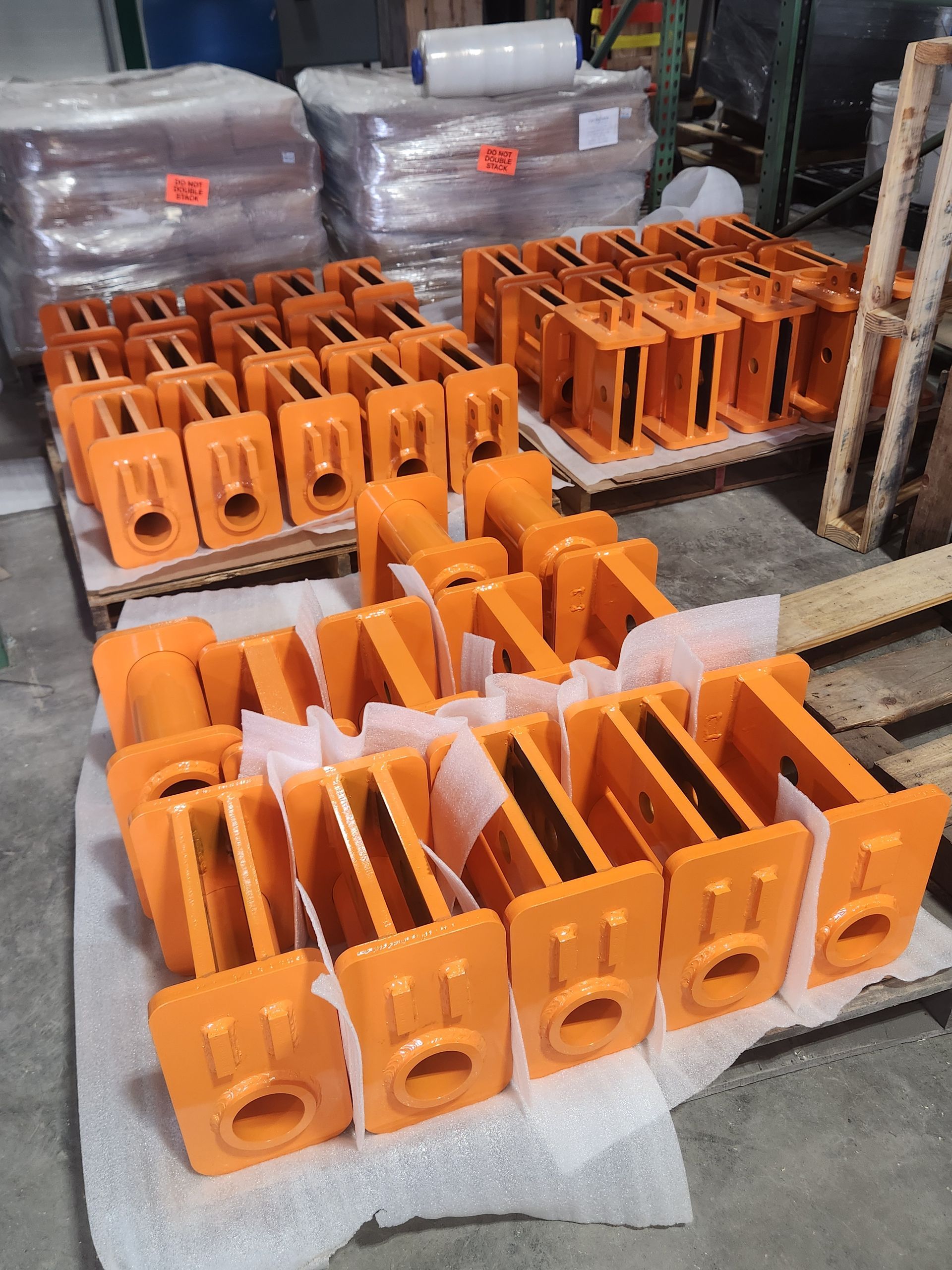Orange metal brackets stacked in rows on a workshop floor, with wrapped bundles in the background.