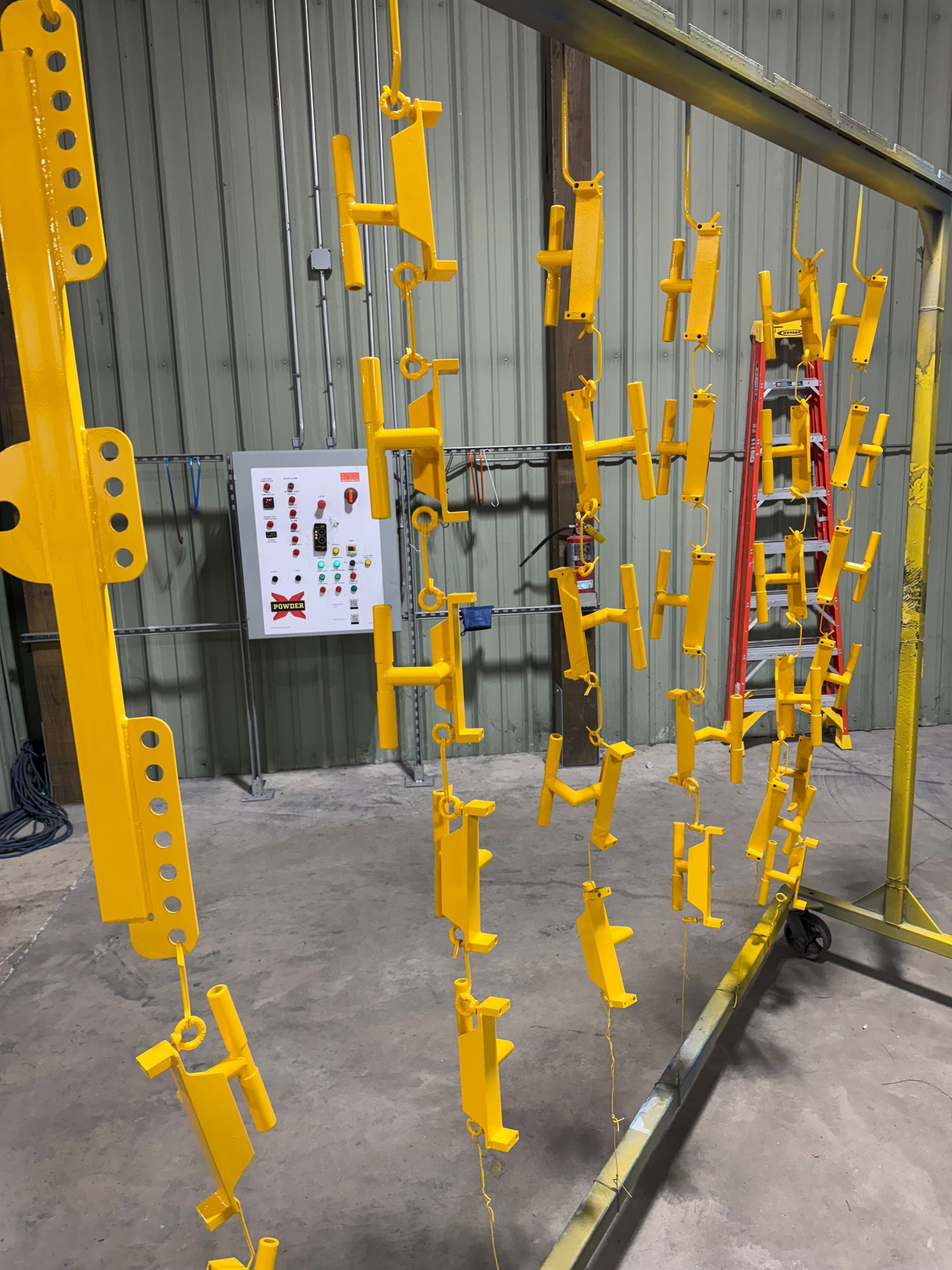 Yellow industrial hanging fixtures displayed in rows against a corrugated metal wall in a workshop.