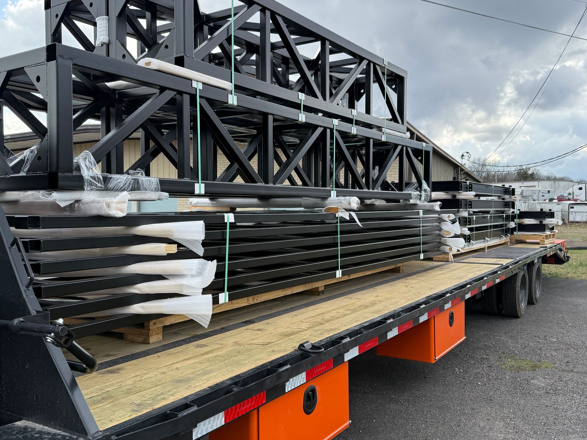 Flatbed trailer loaded with stacked black metal trusses and wrapped materials in an industrial yard