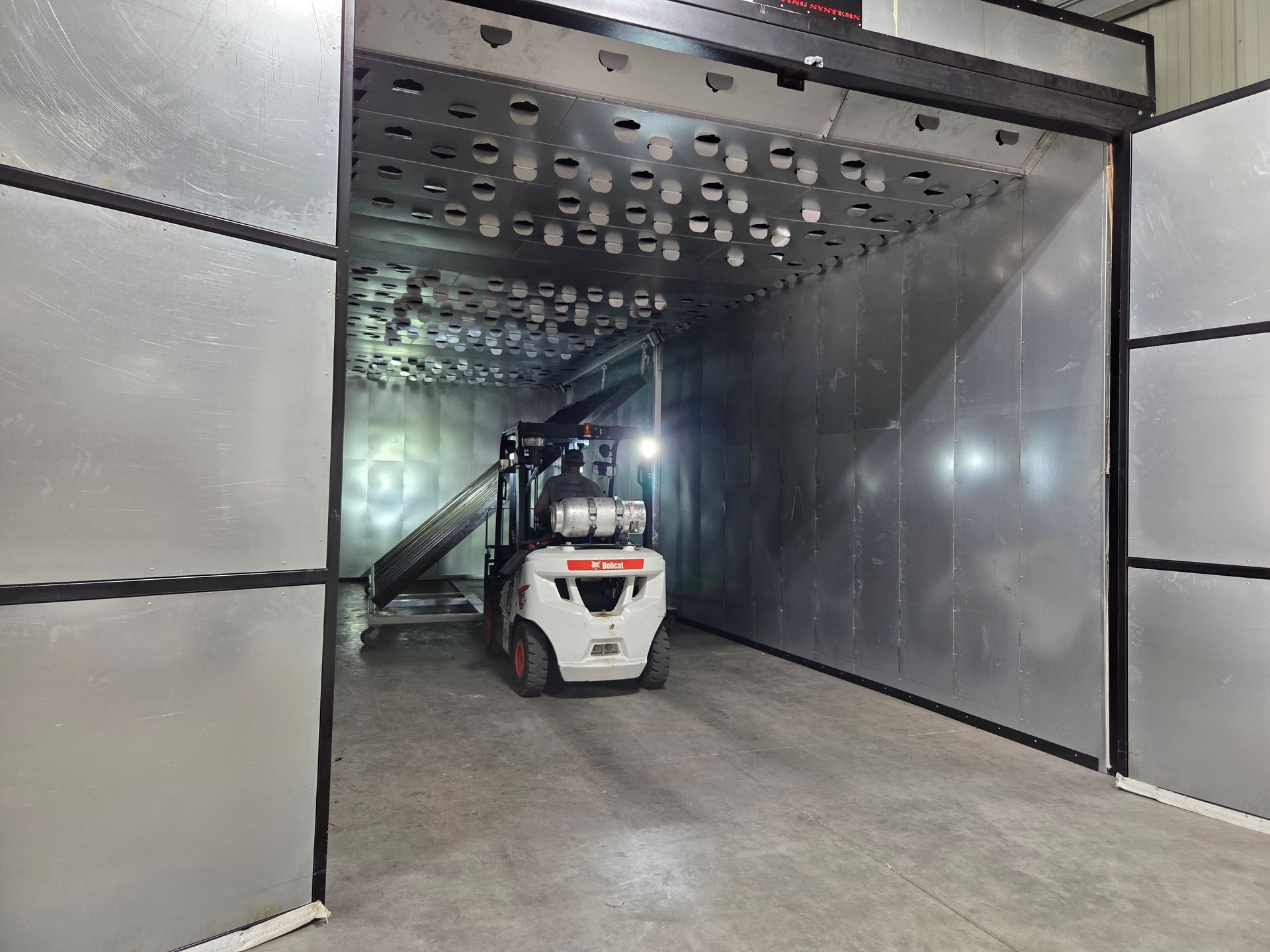 Compact forklift in a metallic industrial corridor with a perforated overhead panel.