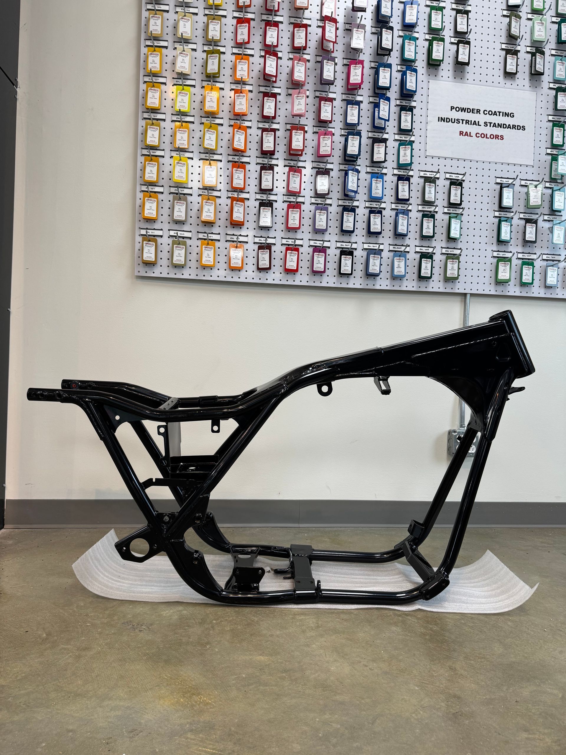 Black motorcycle frame on a concrete floor in a workshop, with a wall display of small parts behind it