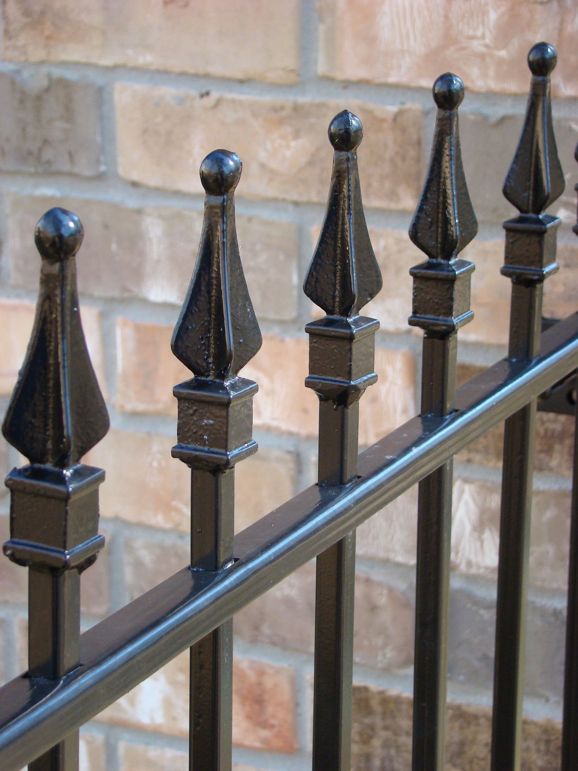 Black wrought-iron fence posts with pointed finials against a tan brick wall.