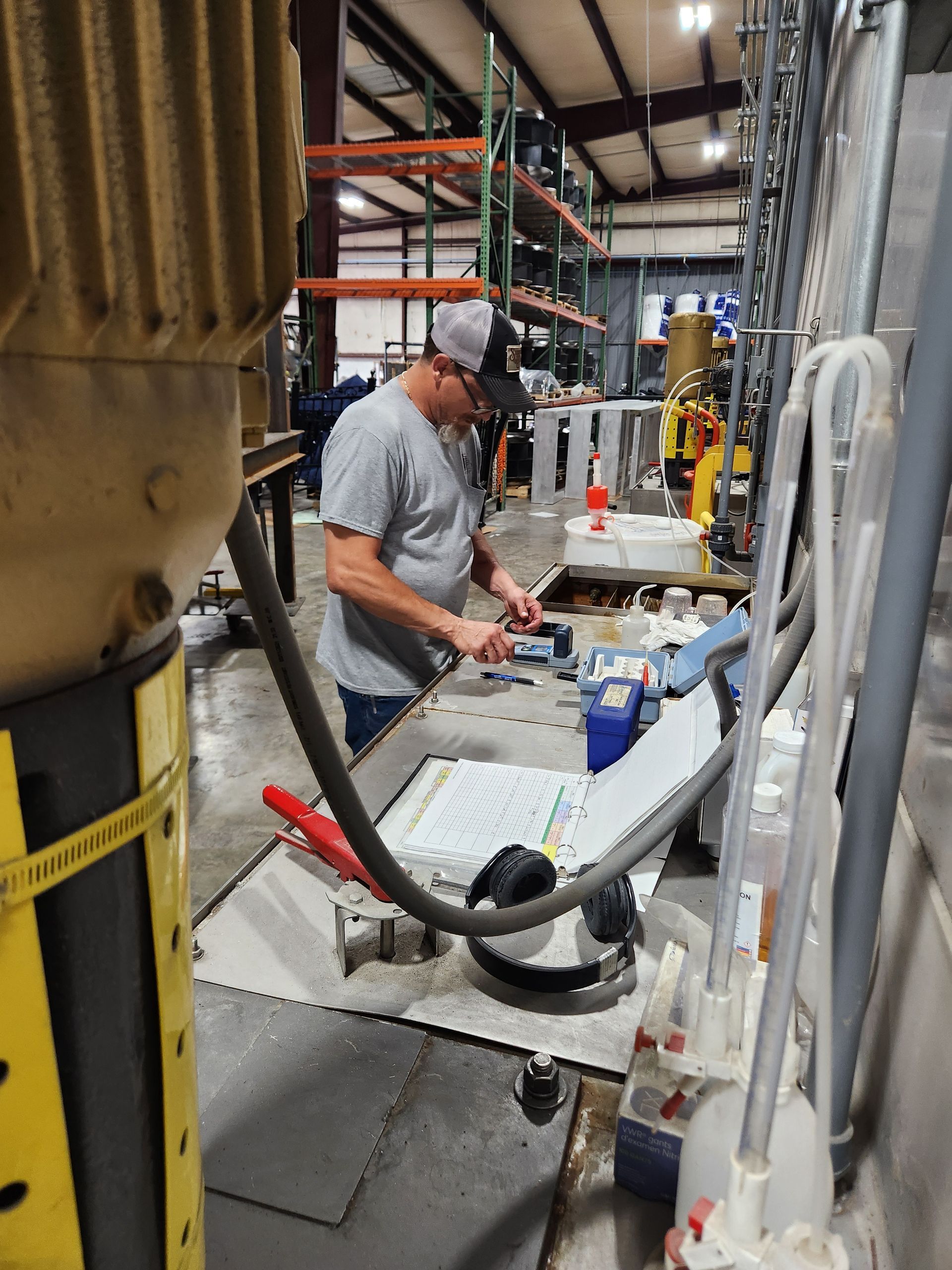 Worker in a factory aisle bends over a workbench, handling equipment amid hoses and industrial machinery.