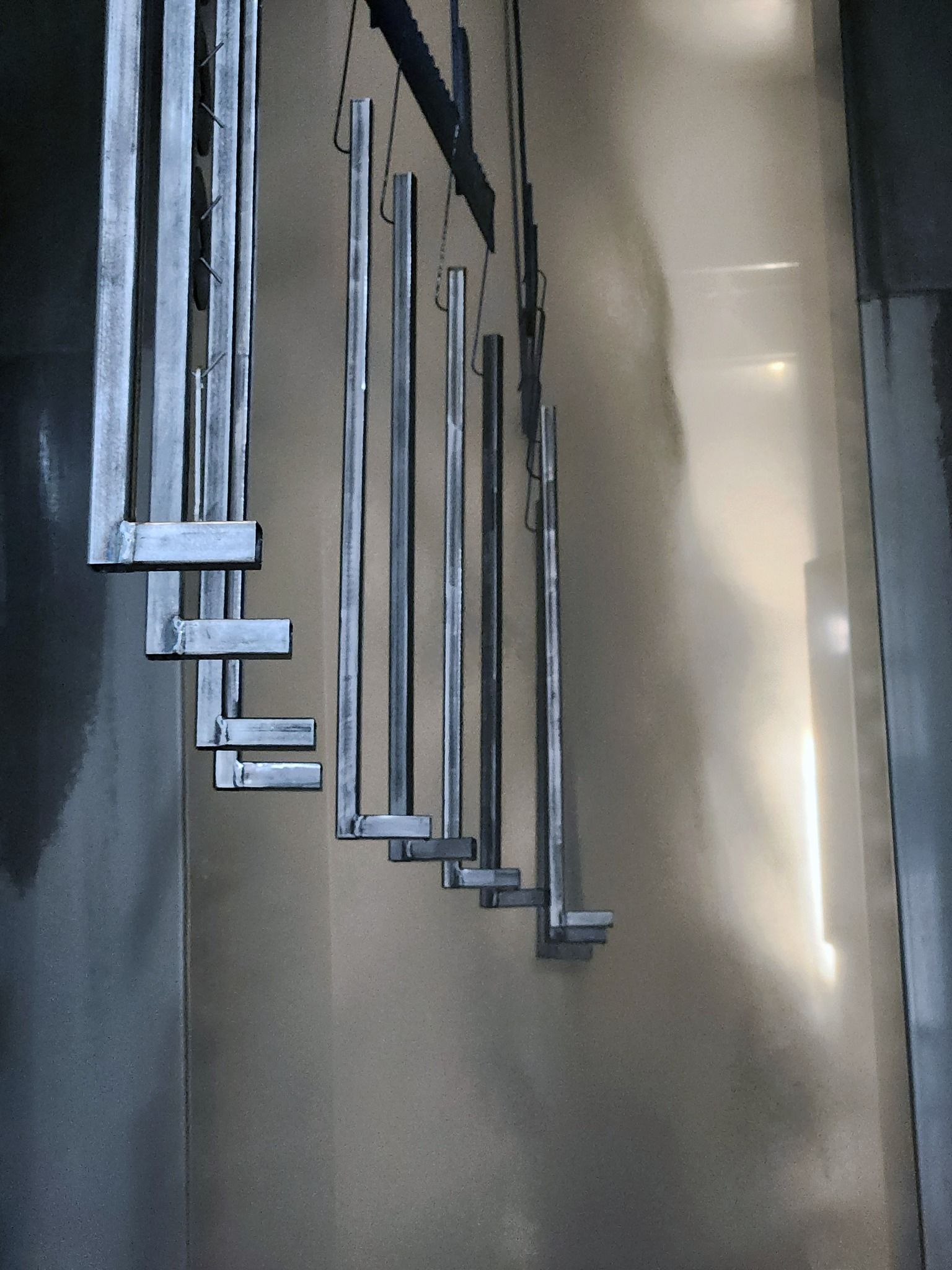 Row of silver metal chime tubes hanging against a gray wall in a narrow indoor corridor
