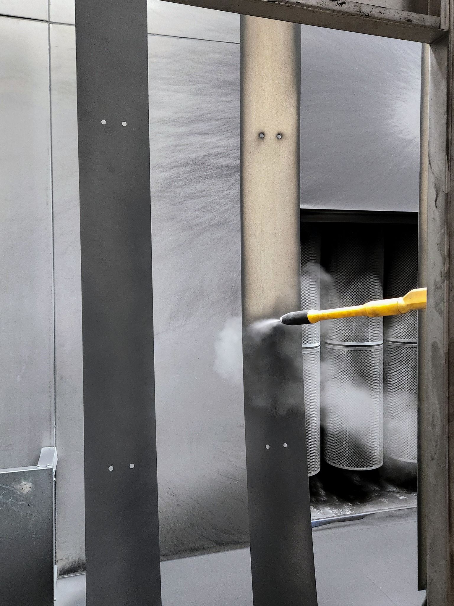 A yellow spray wand mists a gray metal pillar beside a vented wall.