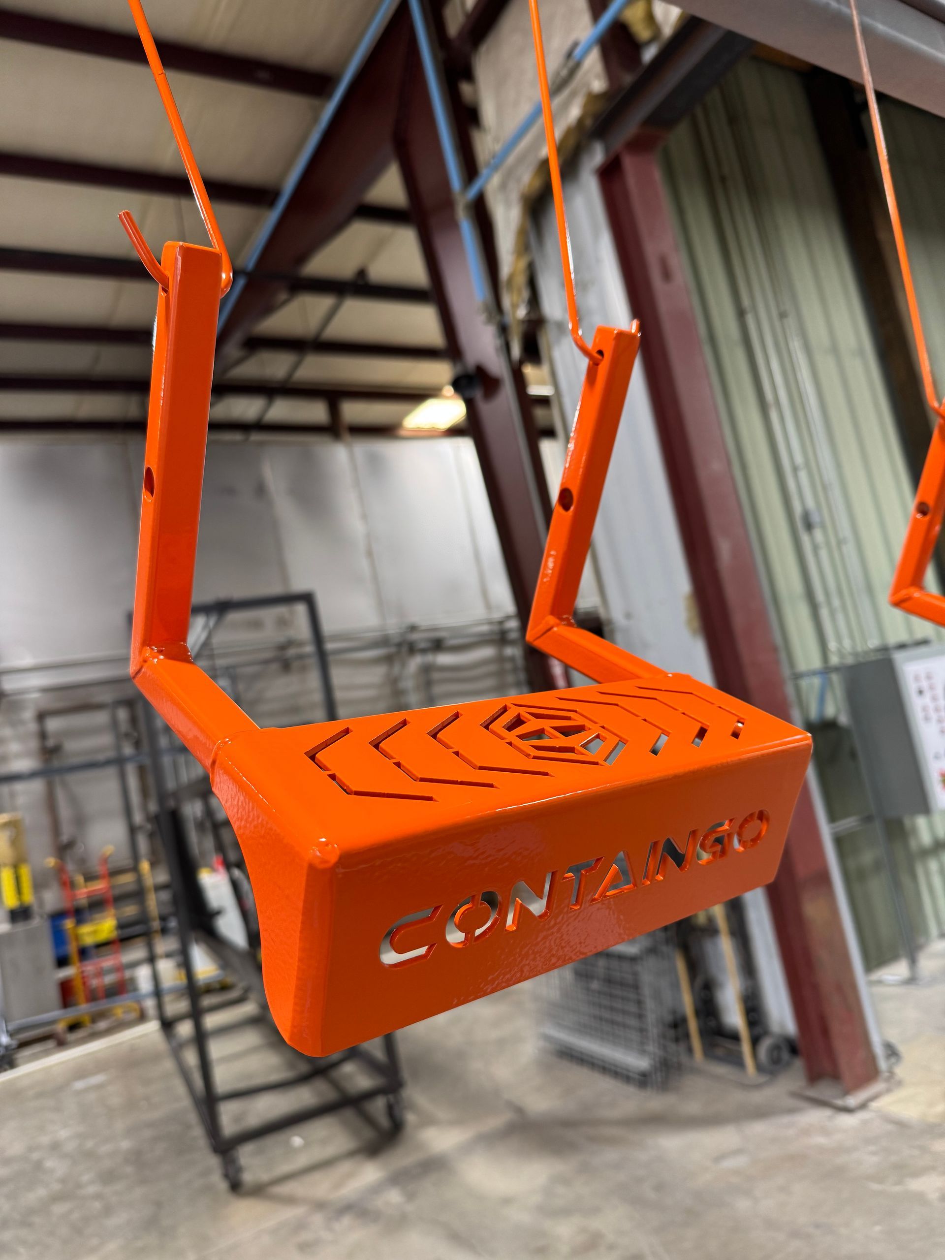 Orange metal lifting fixture hanging in an industrial warehouse, with hook cables attached.