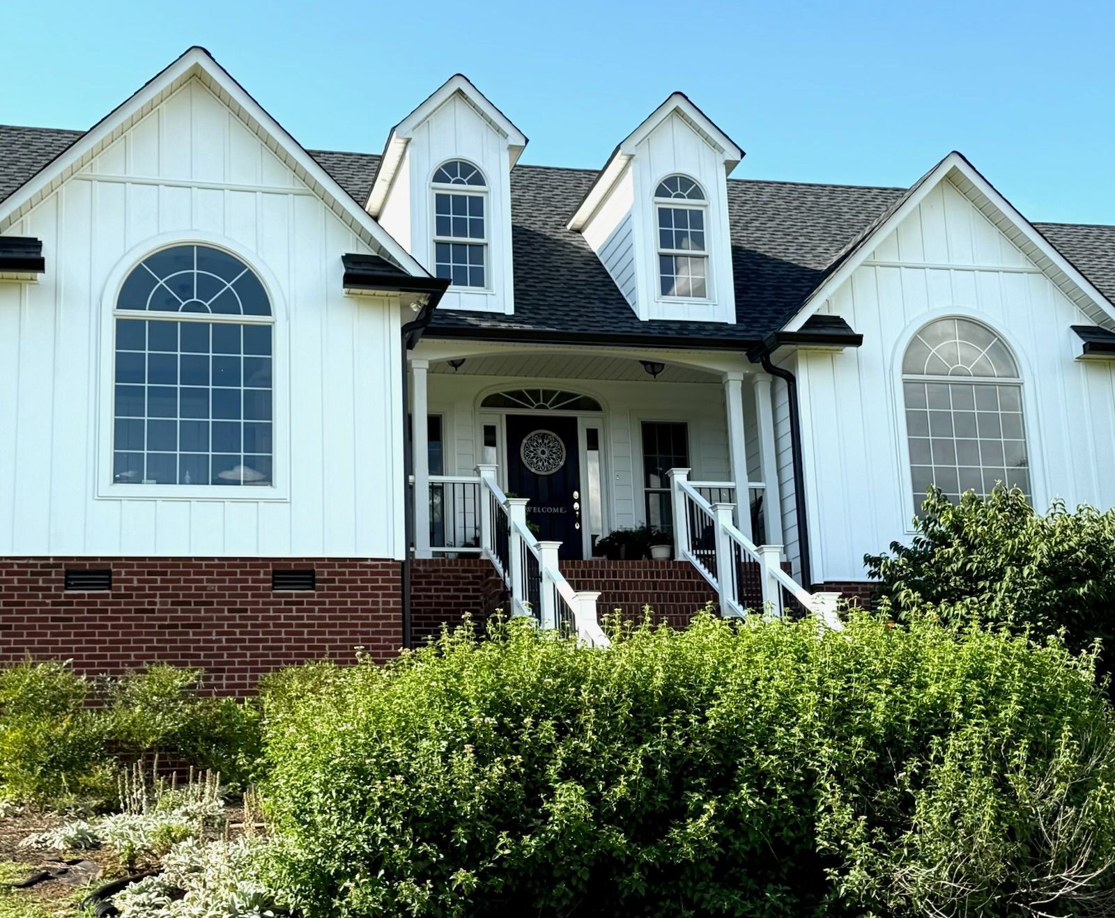 A white house with a black roof is surrounded by bushes