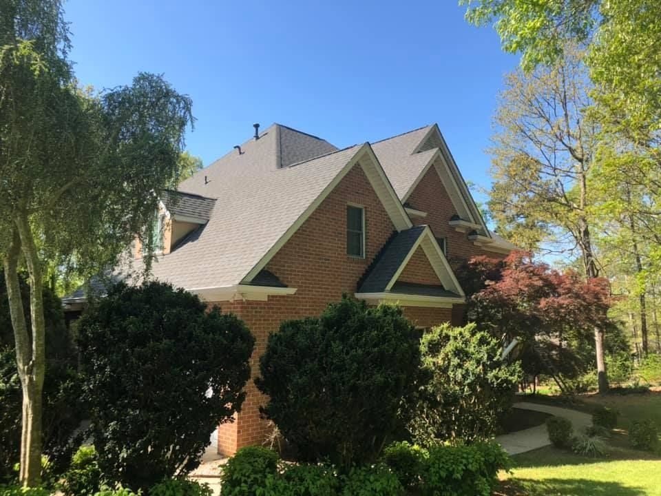 A large brick house with a gray roof is surrounded by trees and bushes.