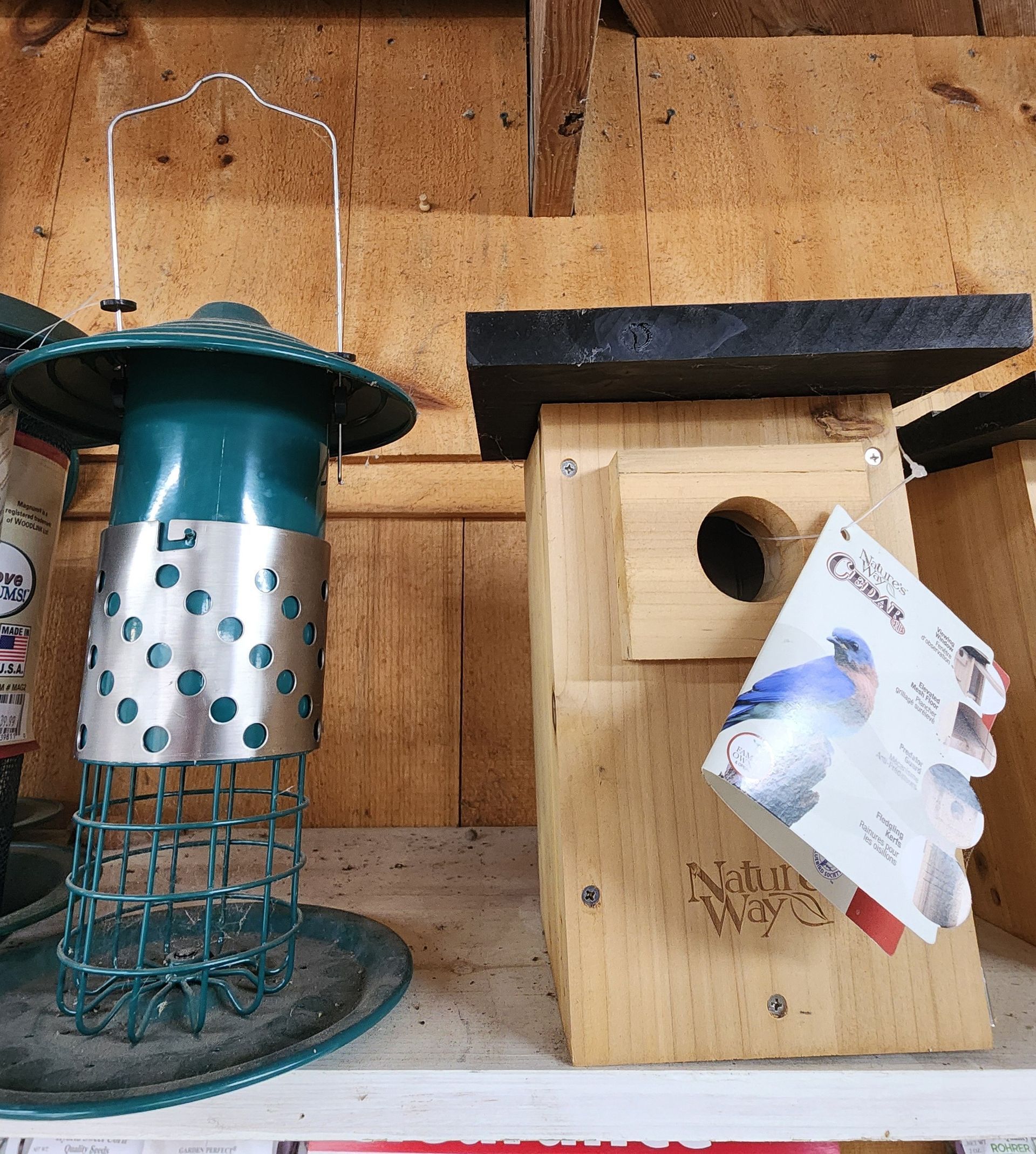 Two bird feeders hanging on a wooden wall, one green-and-silver and one wooden box with a card attached.