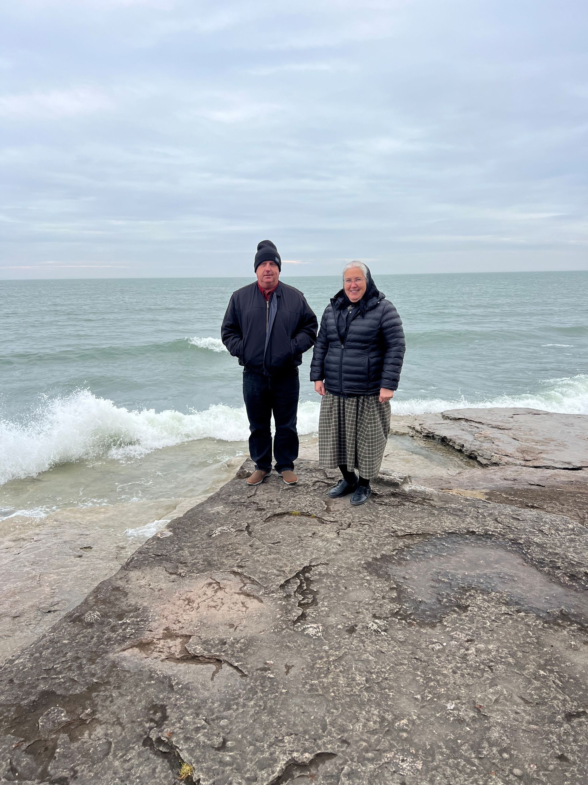 Two people stand on a rocky shore, ocean waves behind them, cloudy sky.