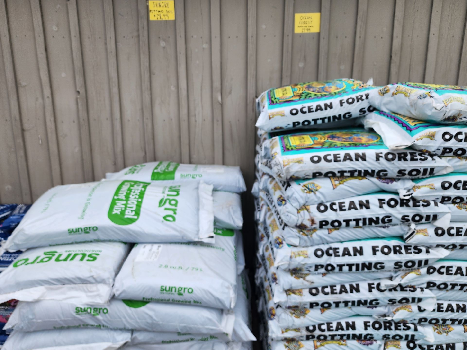 Stacks of white and blue Ocean Forest Potting Soil bags beside a wooden wall outdoors