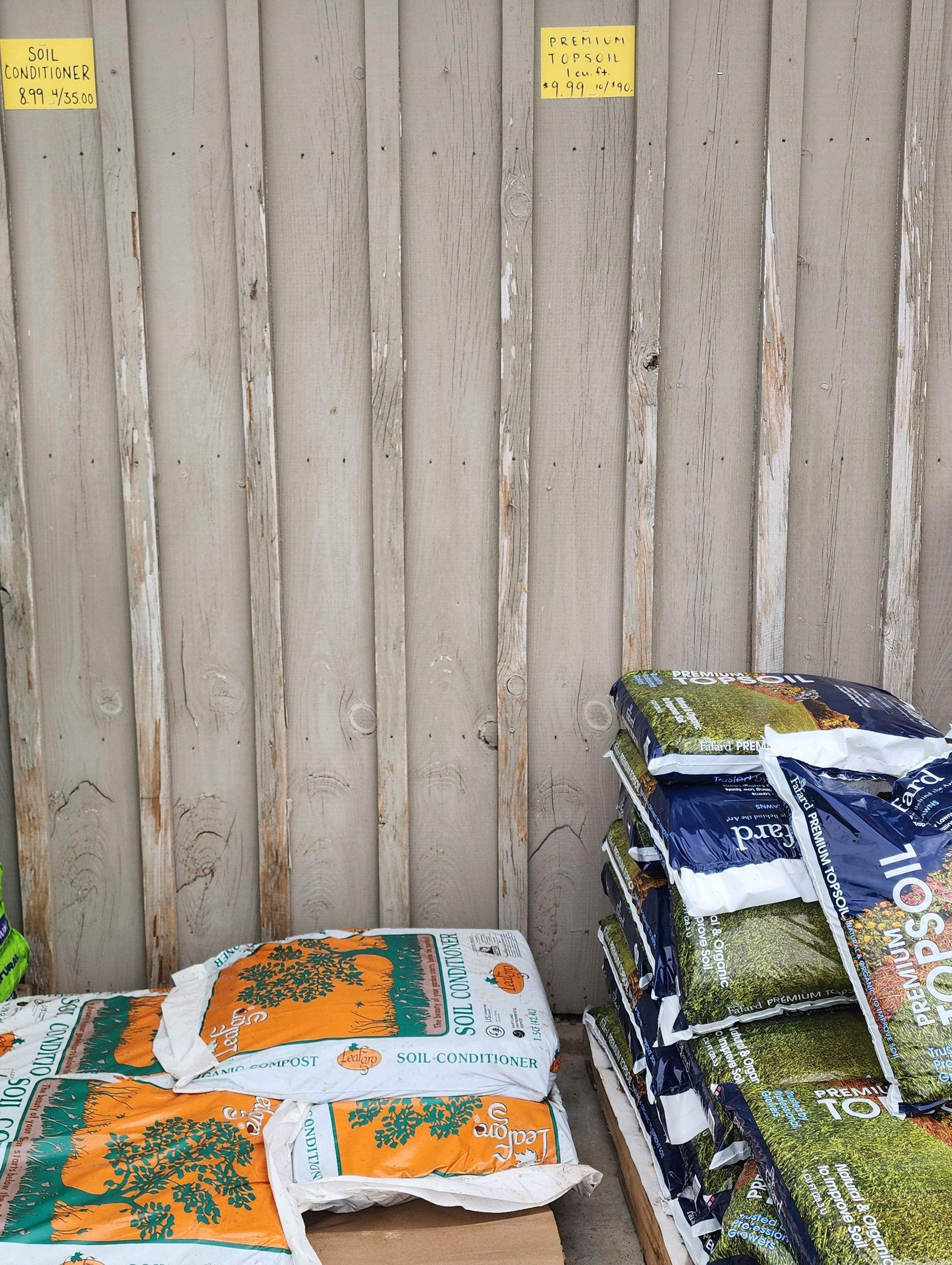 Stacked bags of soil or mulch beside a weathered wooden fence