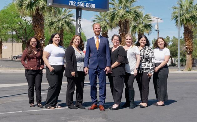 Seven people posing together outdoors in a parking lot under a palm tree sign.