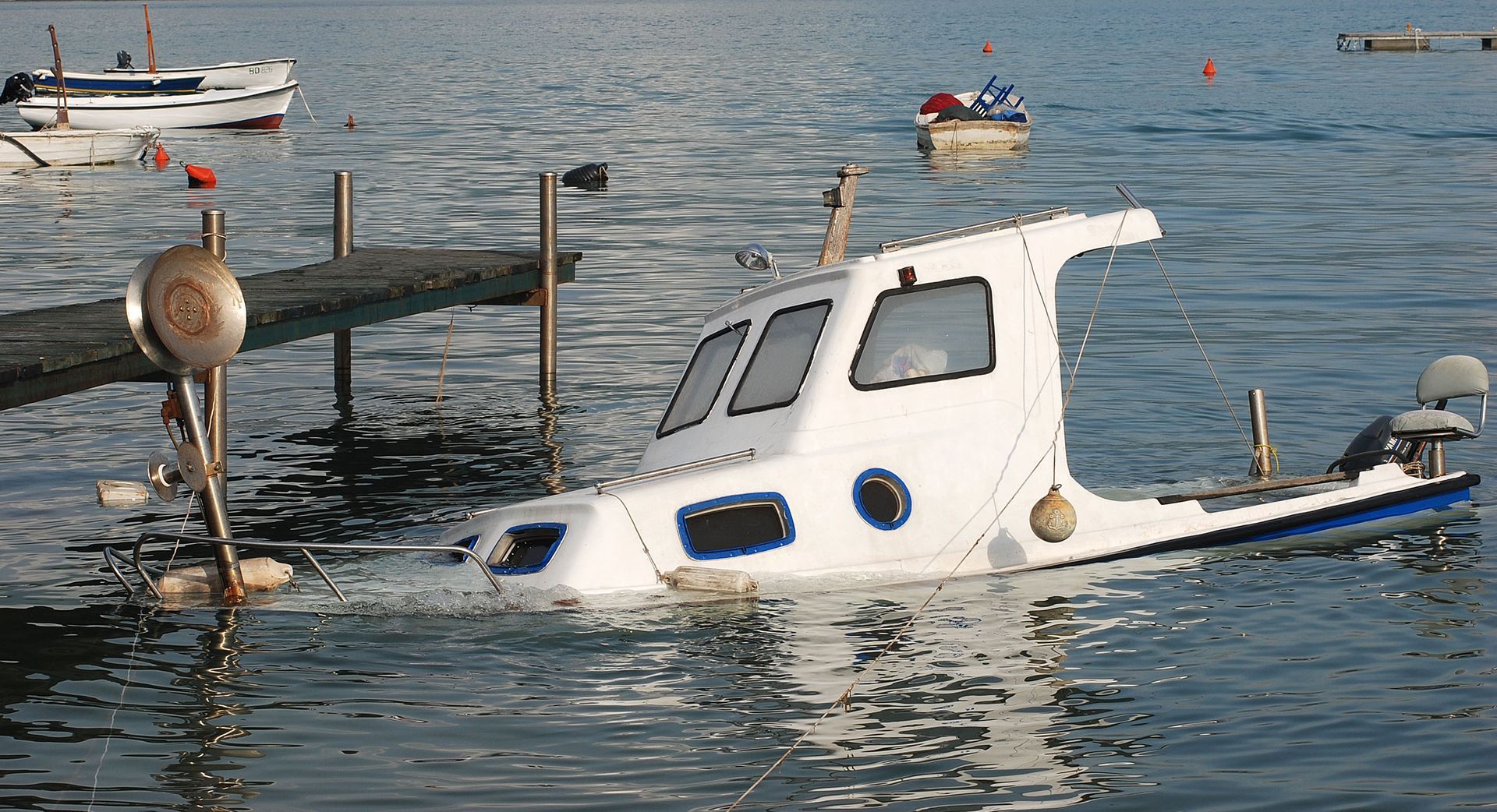 Partially submerged white boat moored by a dock in calm water, with other boats in the background