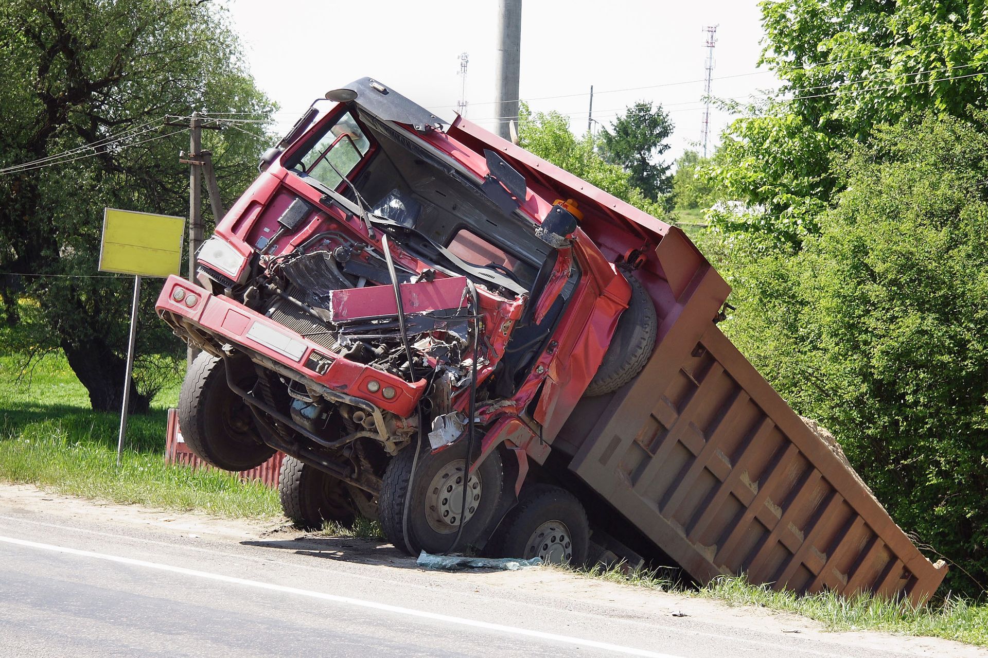 Red dump truck tipped onto its side in a roadside ditch, cab severely damaged.