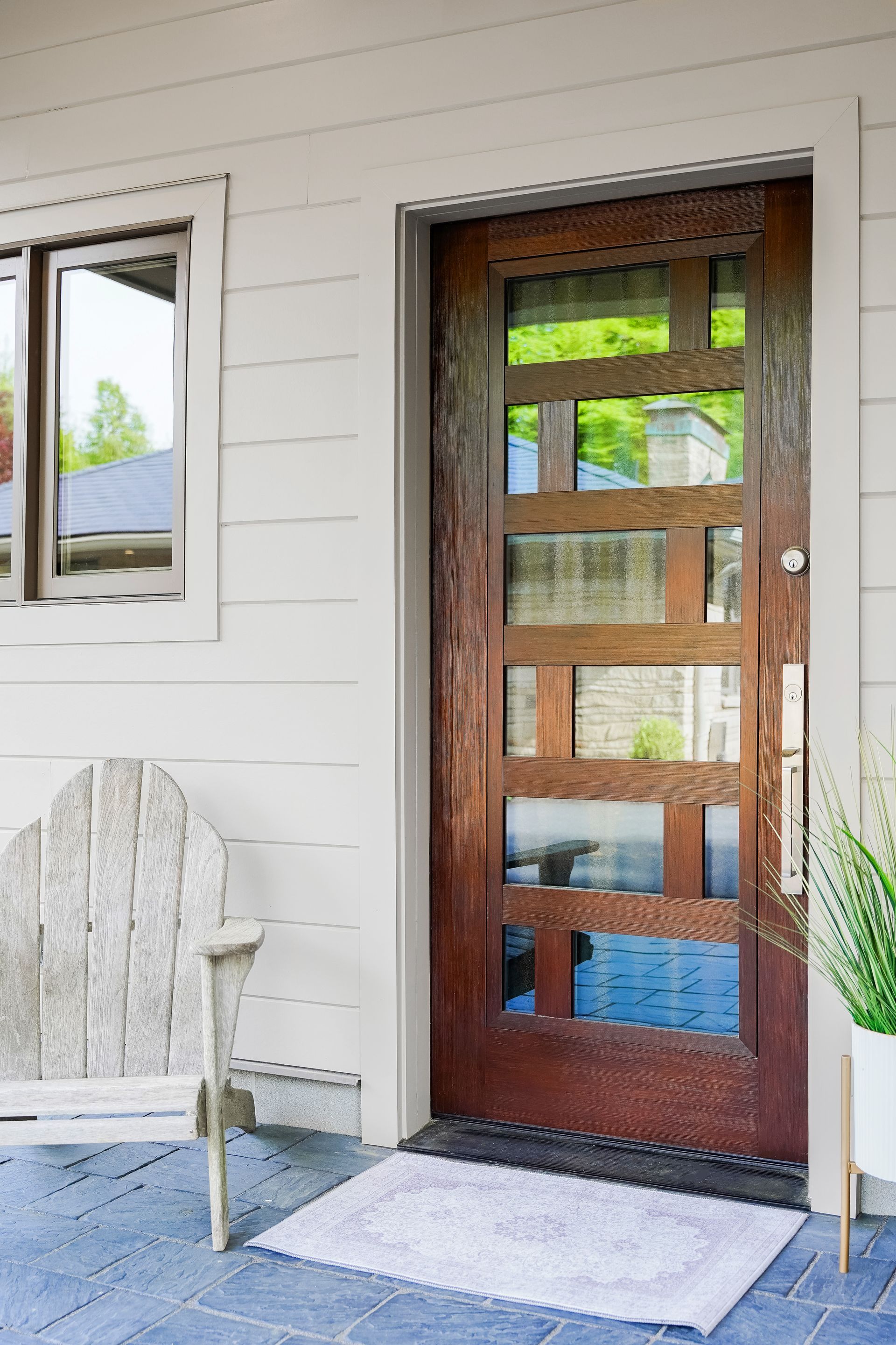 Front porch with wooden glass door, white siding, window, chair, and potted plant.
