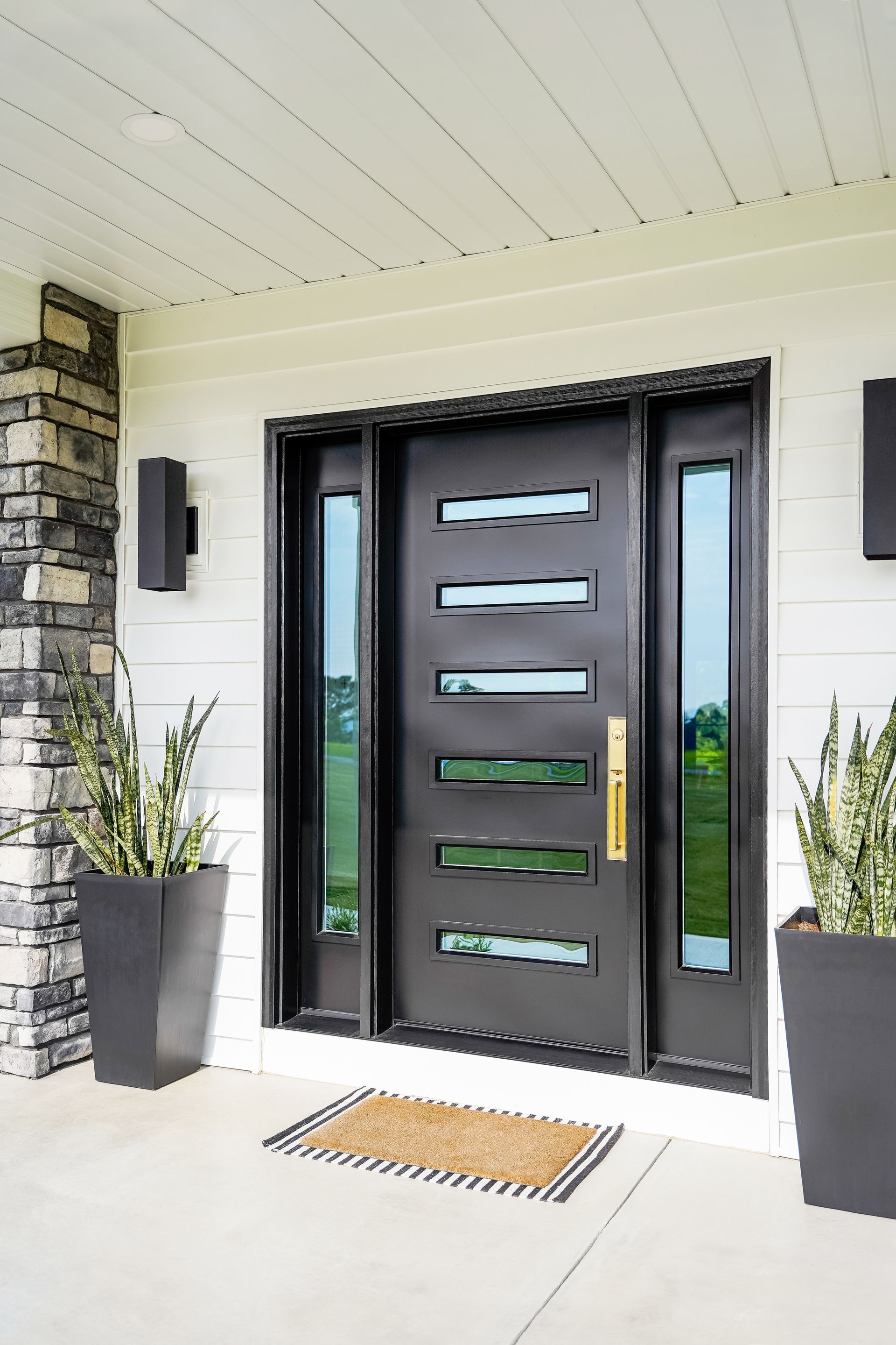 Modern black front door with frosted glass panels, flanked by tall planters on a covered porch