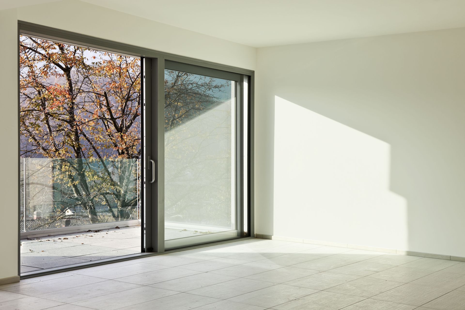 Empty sunlit room with large sliding glass doors and a view of autumn trees outside