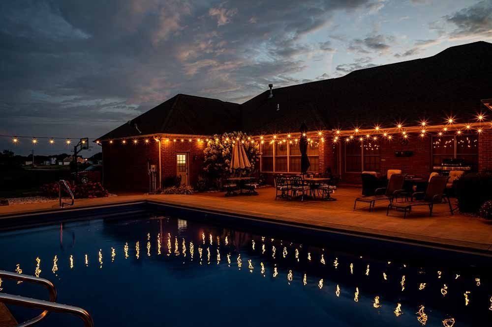 Backyard pool lit by string lights at dusk, reflecting in the water. House in the background.