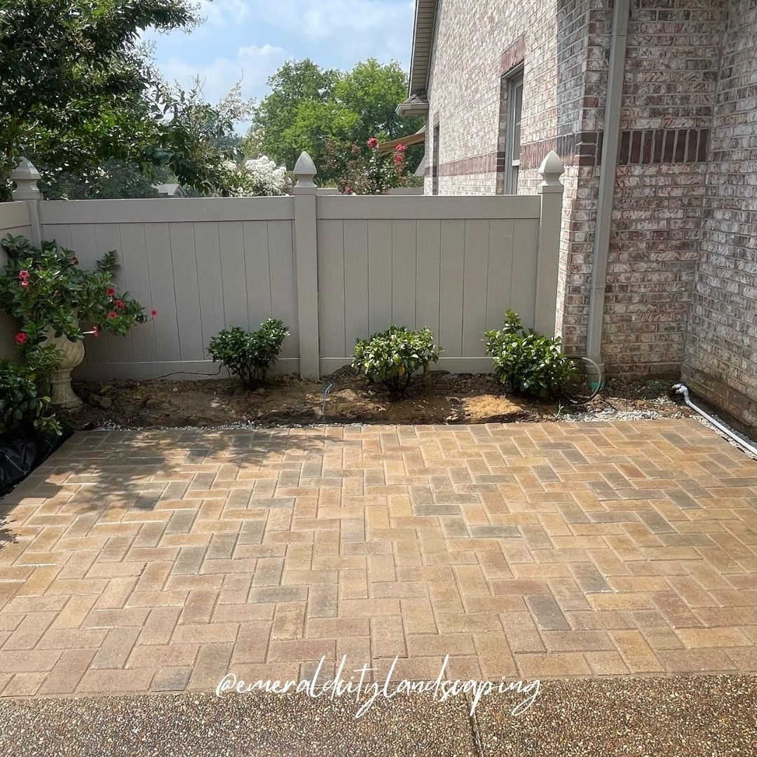 Brick patio with neutral fence and building in the background, with small bushes and a decorative stone.