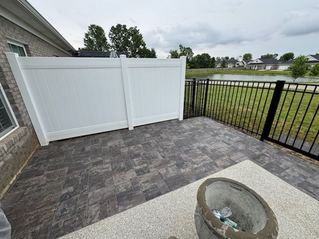 Patio with dark brick pavers and white vinyl fence next to a black iron fence overlooking a pond.
