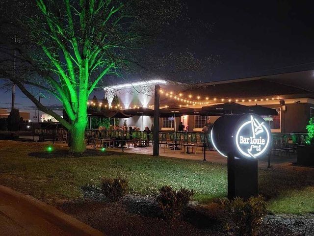 Night view of Blue Suede bar with green-lit tree and sign, patio with lights and tables.