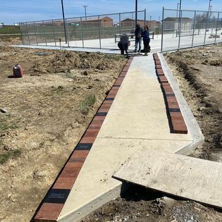 Workers laying brick border along a concrete walkway leading to a fenced area, outdoors.