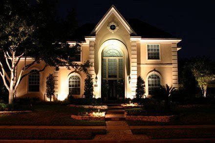 Beige house with warm lighting at night. Lit path leads to the front door. Landscaping with plants.
