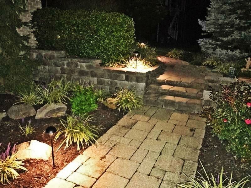 A brick path with steps leads to a garden illuminated by lights at night.