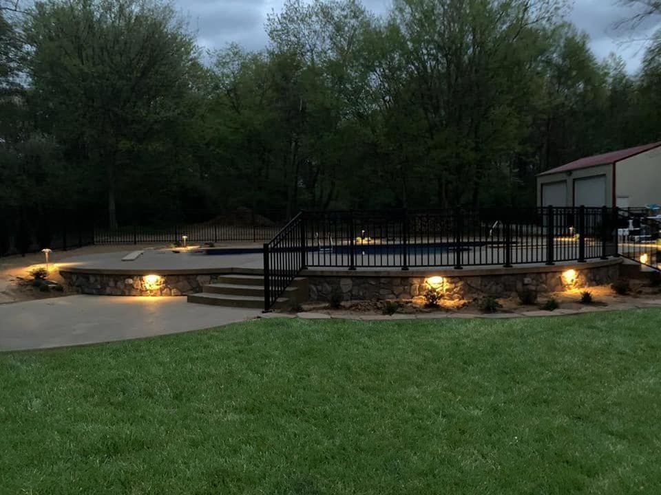 Backyard pool area with lighted stone retaining walls, black fencing, and lush green grass.